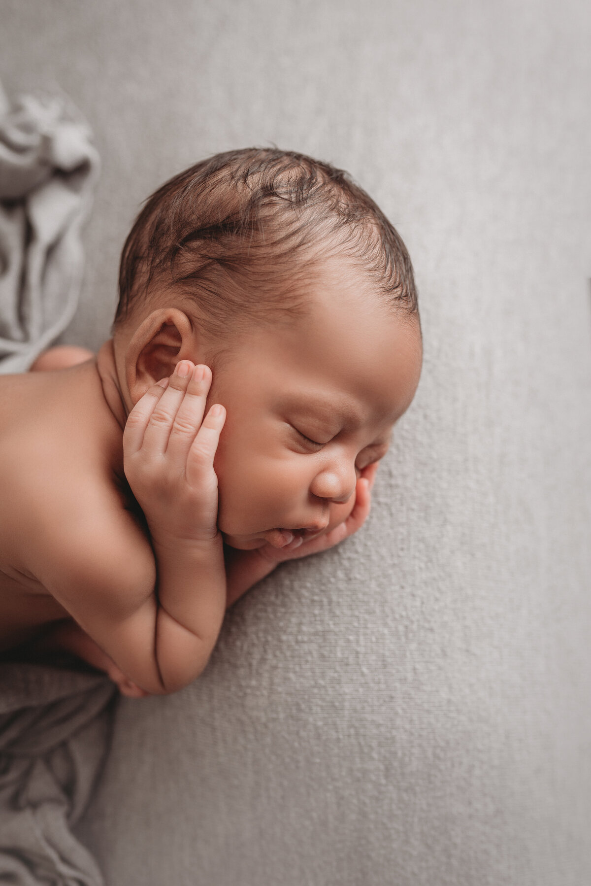 2 week old baby boy asleep on gray fabric backdrop with hands on cheeks