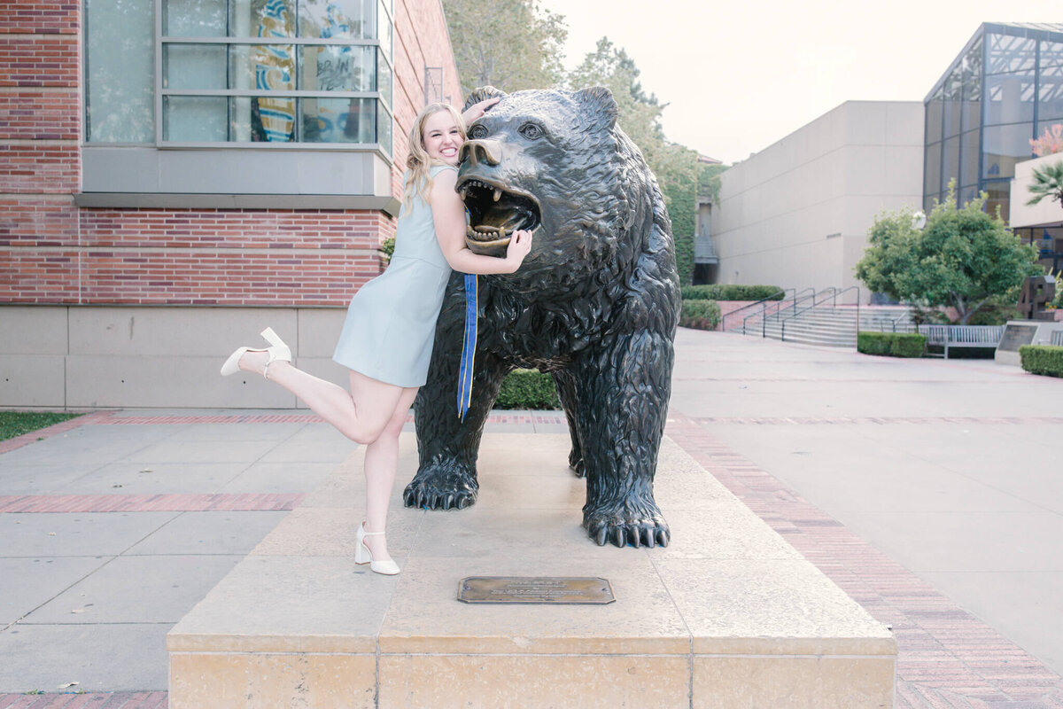 A woman in a light blue dress playfully poses with a large bear statue, lifting one leg. The scene is set on a paved area in front of a building with trees and benches nearby.