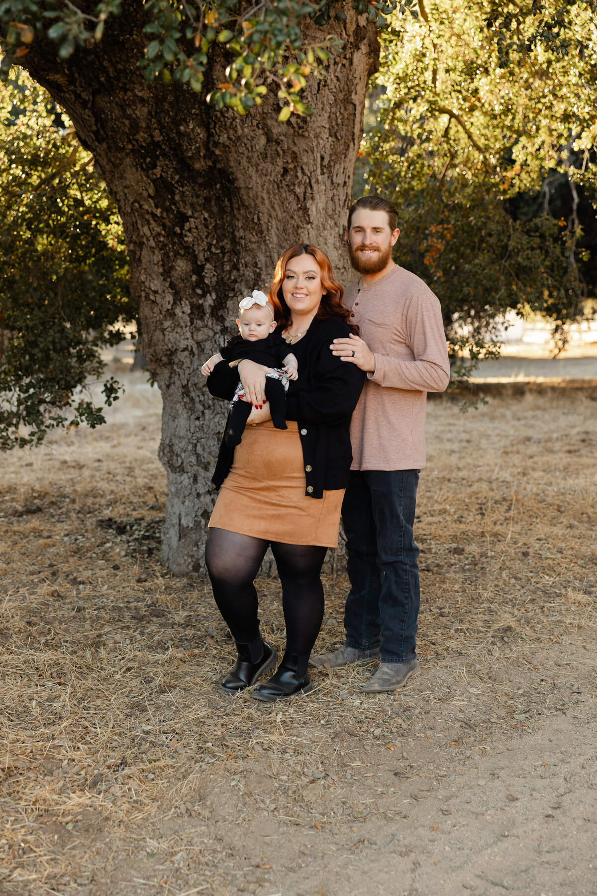 Parents holding their baby under a large tree at sunset, dressed in warm neutral tones. Photographed by Yucaipa family photographer Kaitlyn Dawn Photography.