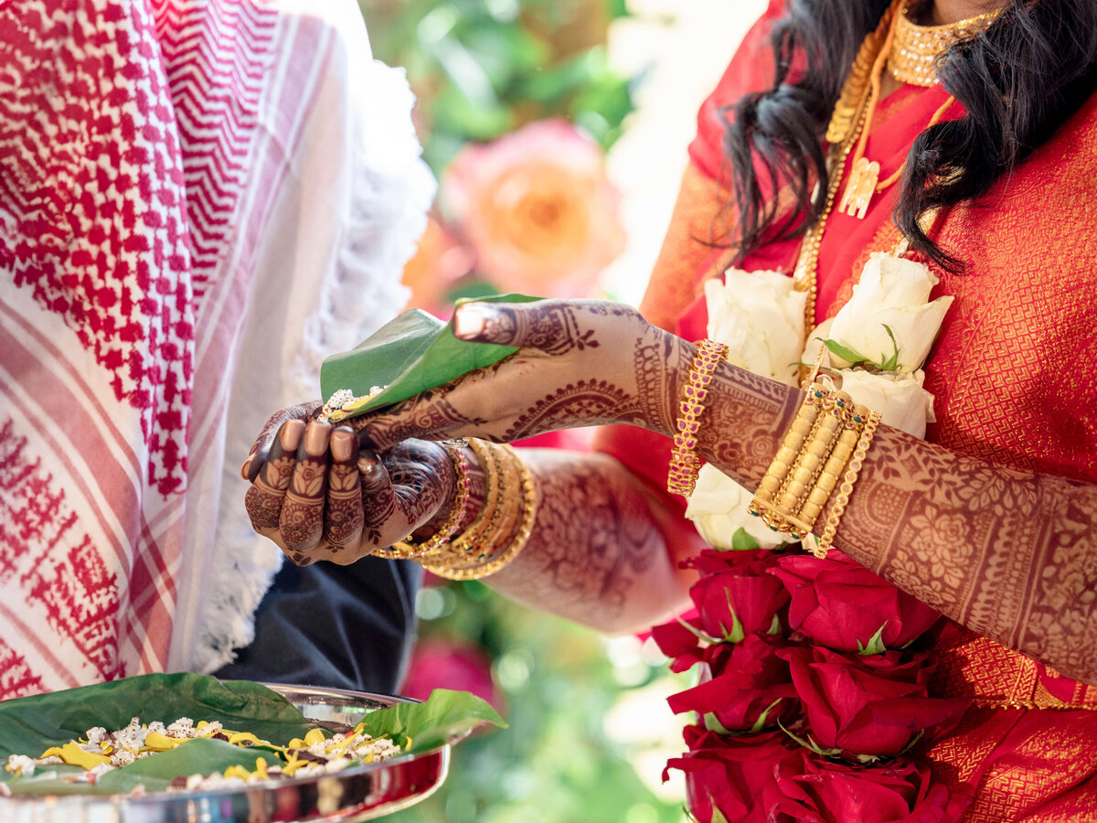 A close-up of a bride in traditional attire holding a green leaf with henna-adorned hands, wearing gold jewelry and a red sari with white and red floral garlands. Another person in traditional dress is partially visible.