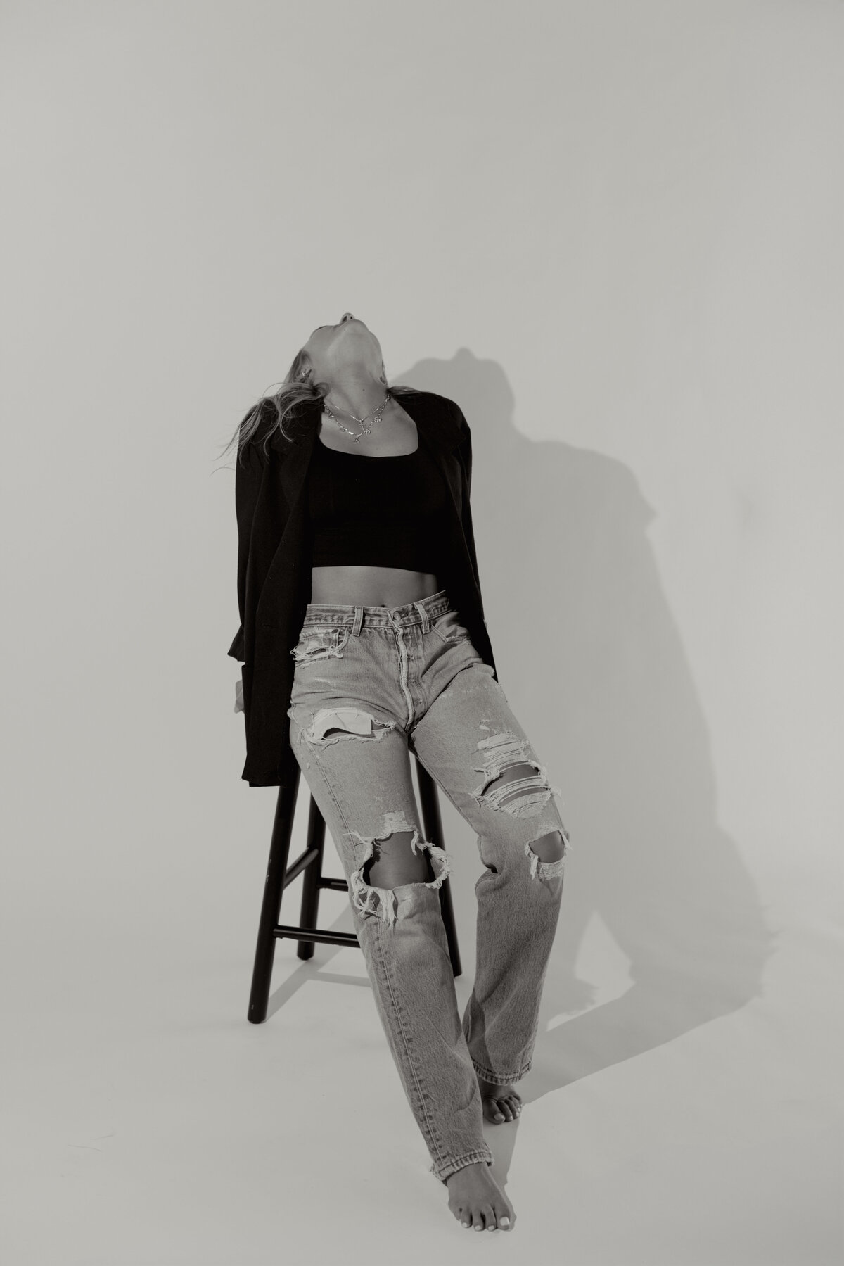 A dramatic black and white studio portrait of a woman sitting on a stool in ripped jeans and a blazer, head thrown back for an expressive, editorial pose. Captured by Dani Lacey in her signature cinematic style in Greenville,SC