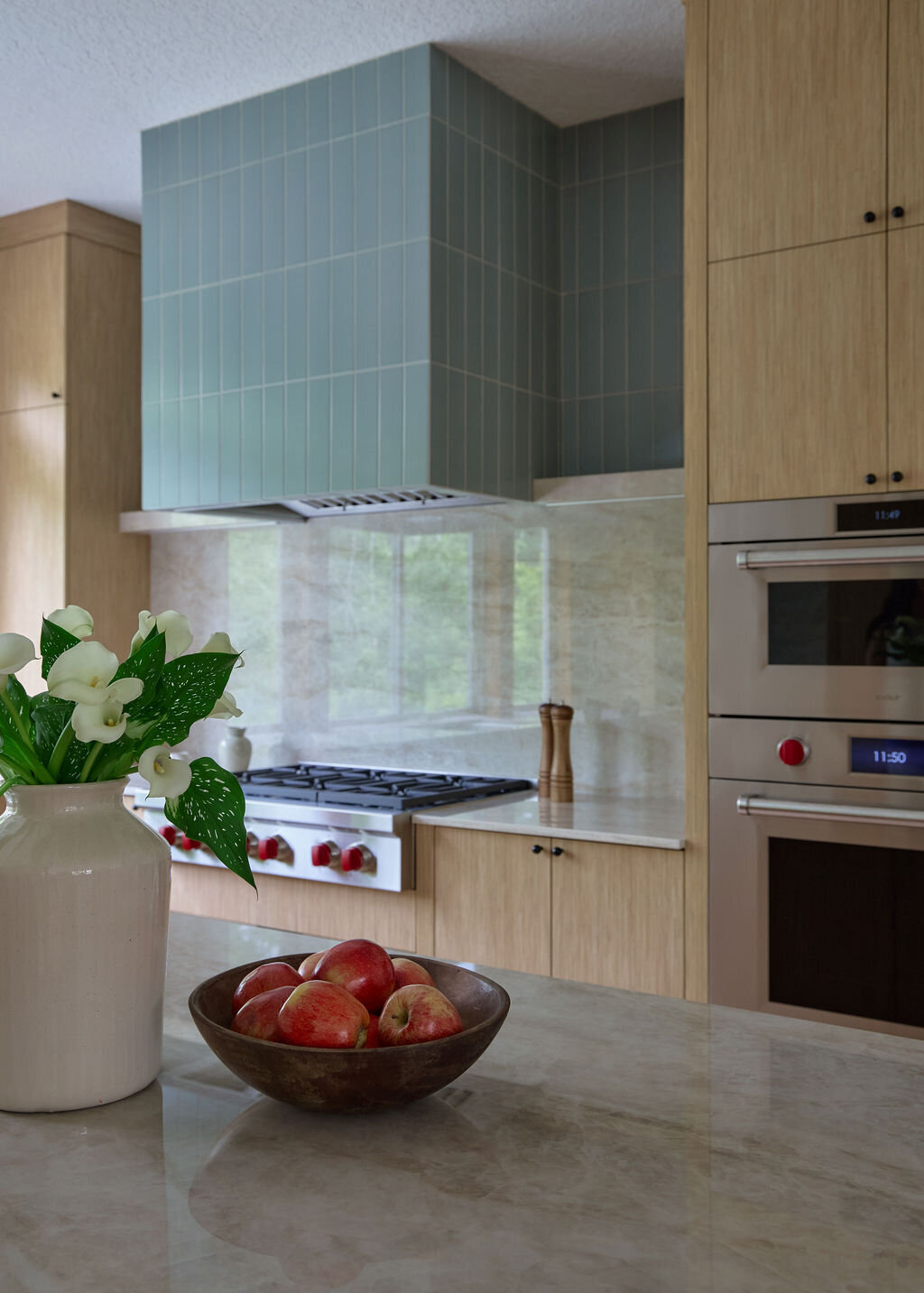 Kitchen with white oak cabinets, and slim simple black hardware. Taj mahal quartzite countertops that run up the backsplash to a quartz shelf with cookbooks and plants on it. Green subway tile backsplash above the shelf to the ceiling. Two sconces above the shelf that are modern, white and gold.
