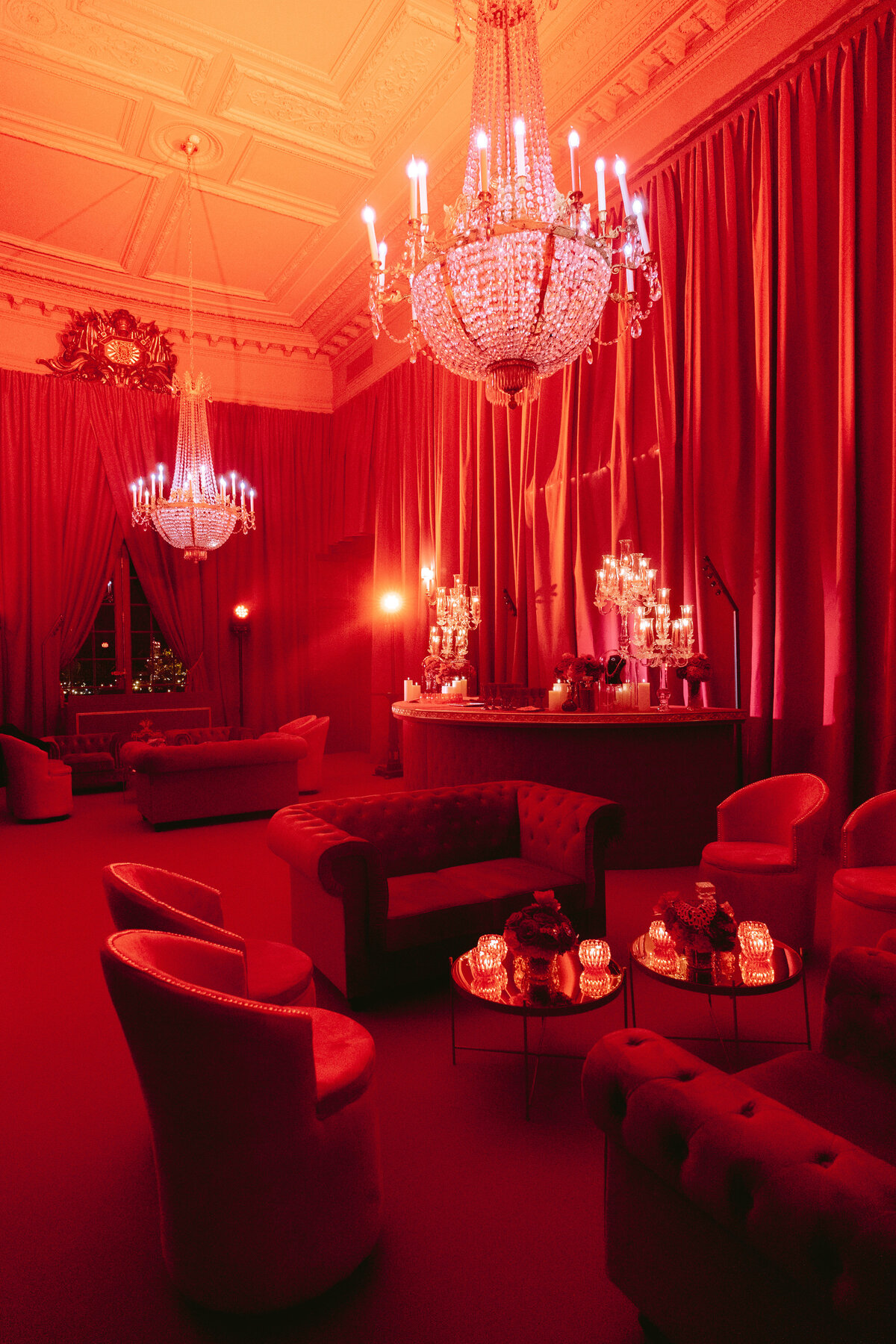 The photo was taken at the opulent Shangri La Hotel & Ritz Paris Hotel in Paris, France, by the talented photographer Matteo Coltro. The lavish room is bathed in a warm red light, accentuating its grandeur. It features two exquisite chandeliers that illuminate the space, plush tufted sofas, and cozy armchairs inviting you to unwind in style. Decorative candles and lights grace the tables, adding to the room's luxurious and intimate ambiance. Additionally, Zen Film Works, a team of skilled videographers, captured the beauty of this setting through their lens to create stunning visual content.