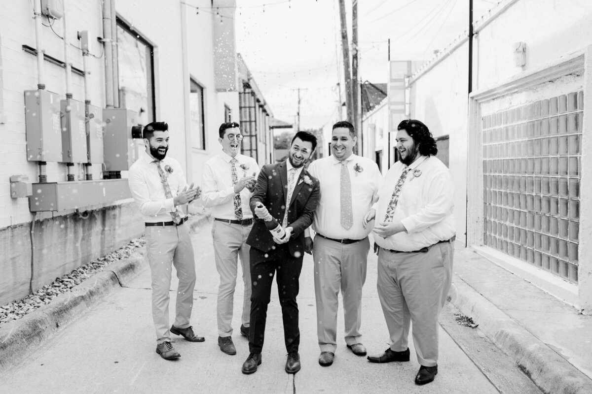 Black and white action shot of a groom spraying a bottle of champagne in an alley while his groomsmen cheer him on.  The groom is in the middle of the group and is wearing a dark suit with a boutonniere. Two groomsmen are on either side of him and wear dress shirts, dress pants, ties, and boutonnieres.