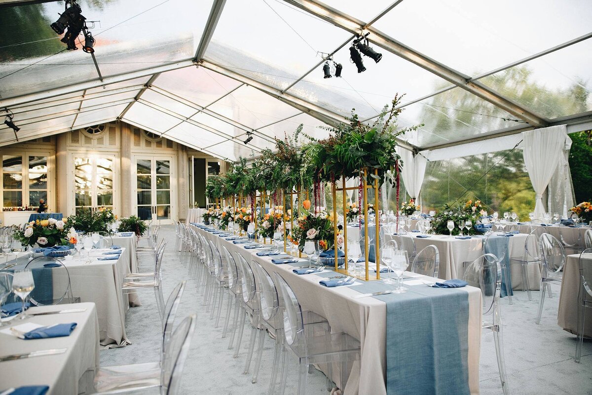 Inside a clear topped tent at Cheekwood are long Downton Abbey style tables set for a wedding. The tables are covered in white table cloths with wide textured blue table runners and are surrounded by clear acrylic chairs. The white plates are topped with blue linen napkins and set with gold flatware and stemmed crystal glasses. At the center of the tables are tall floral centerpieces on high gold stands. The flowers are yellow and orange. There are high and low floral arrangements on each table.