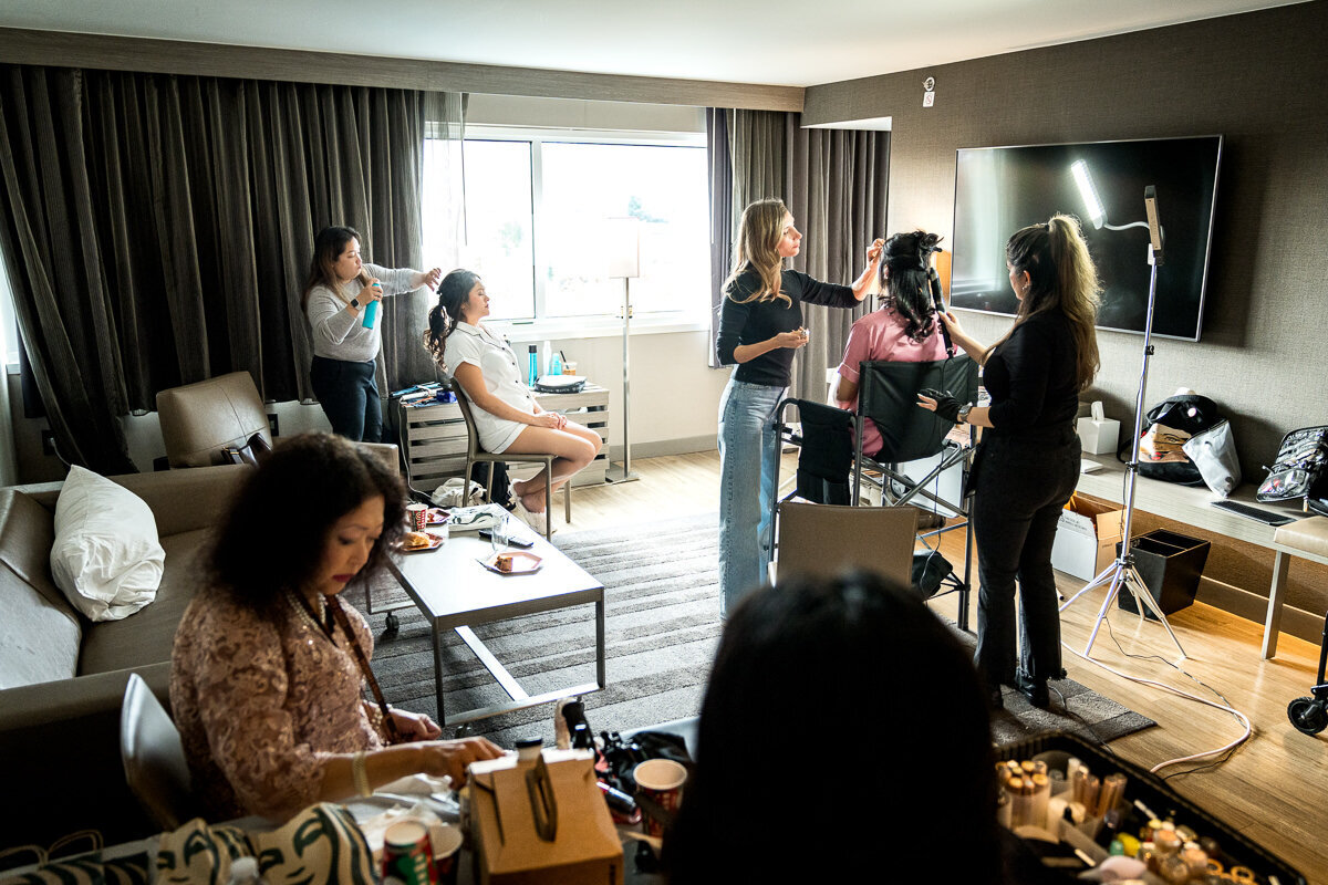 The bride and her bridesmaids share laughter and excitement while getting their hair and makeup done in the elegant hotel room at AC Hotel Pleasanton, capturing the joyful moments leading up to the wedding.