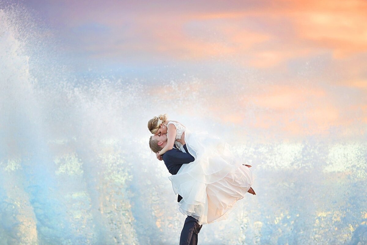 Groom lifts his bride wearing a fluffy wedding gown as a powerful wave crashes behind them after their engagement portraits on Maui