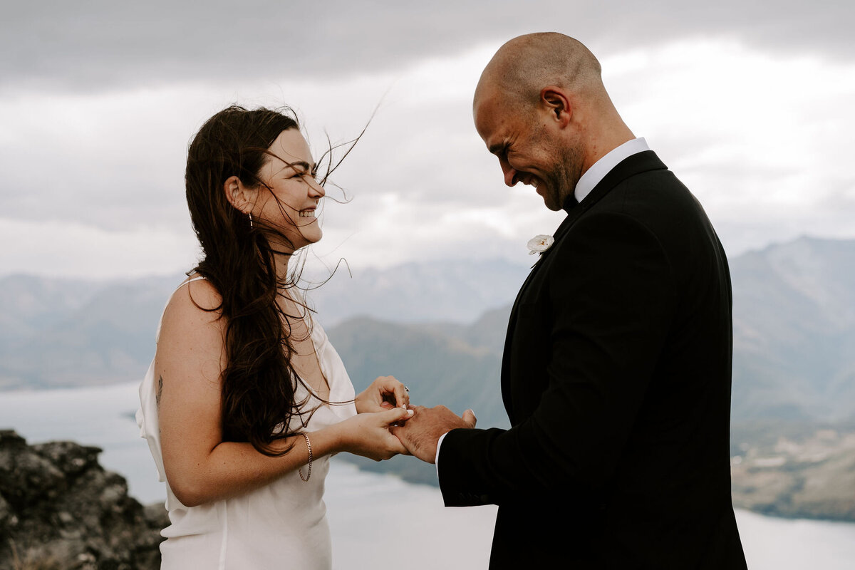 The Lovers Elopement Co - bride and groom exchange rings on top of mountain with lake below, heli wedding