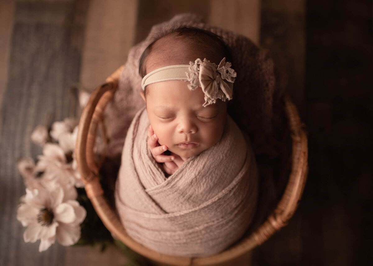 Close-up, aerial image. Baby sleeping in basket in a creamy blush wrap with a cream headband. Flowers are placed beside the basket.