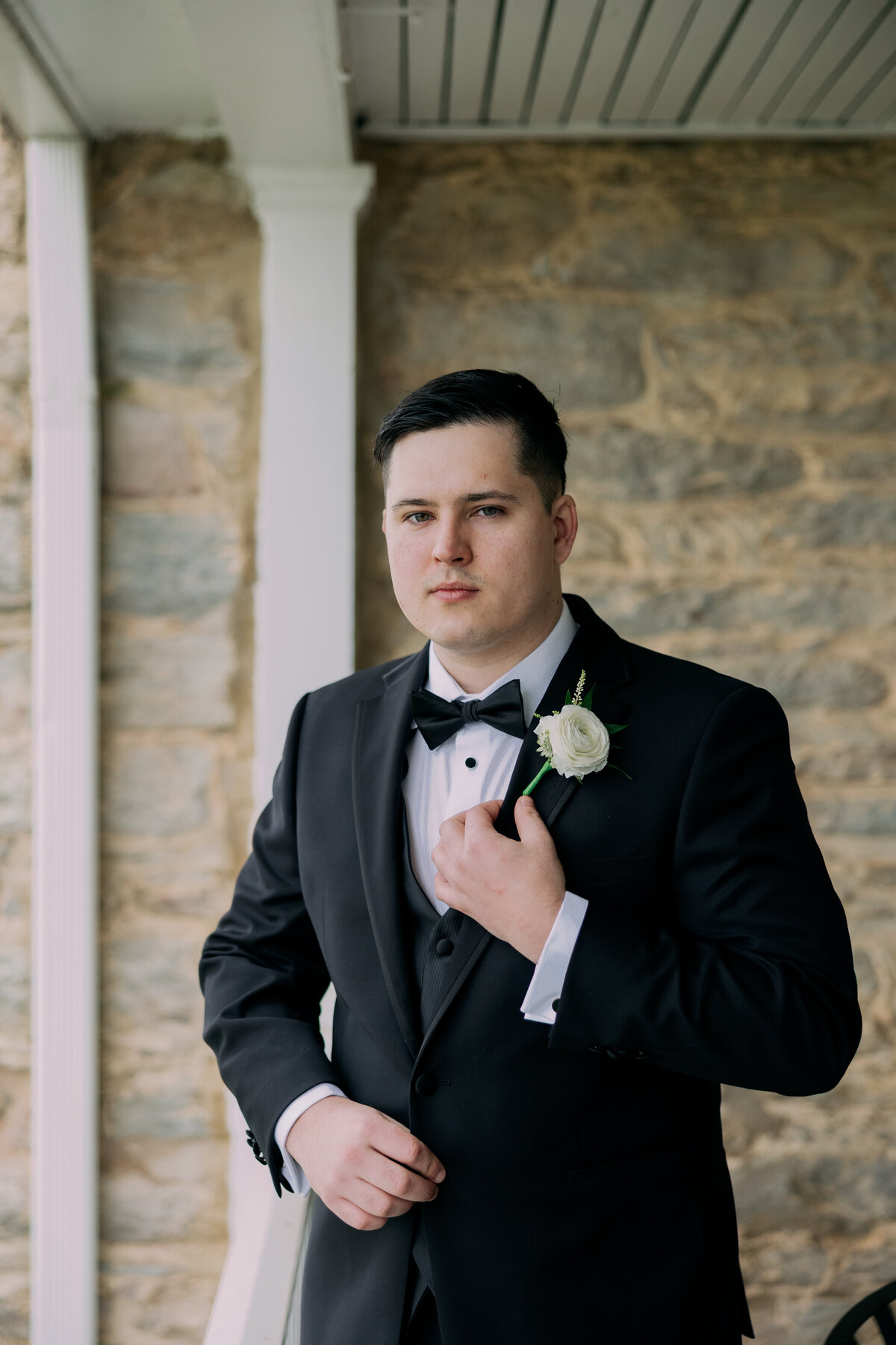 Groom adjusting tuxedo jacket during spring portraits outside Springfield Manor’s stone walls.