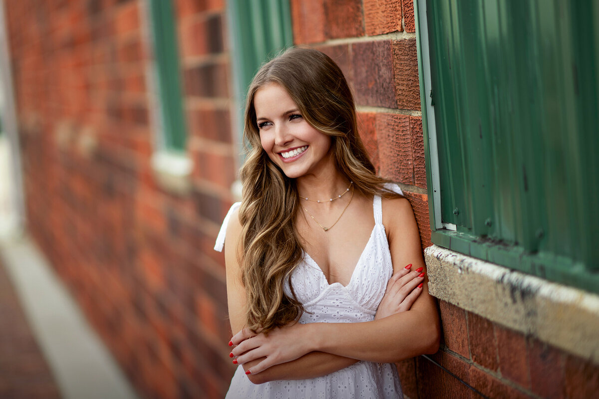 An image from a Pella Senior Photographer A high school senior leans on a wall while smiling in a white dress with arms crossed