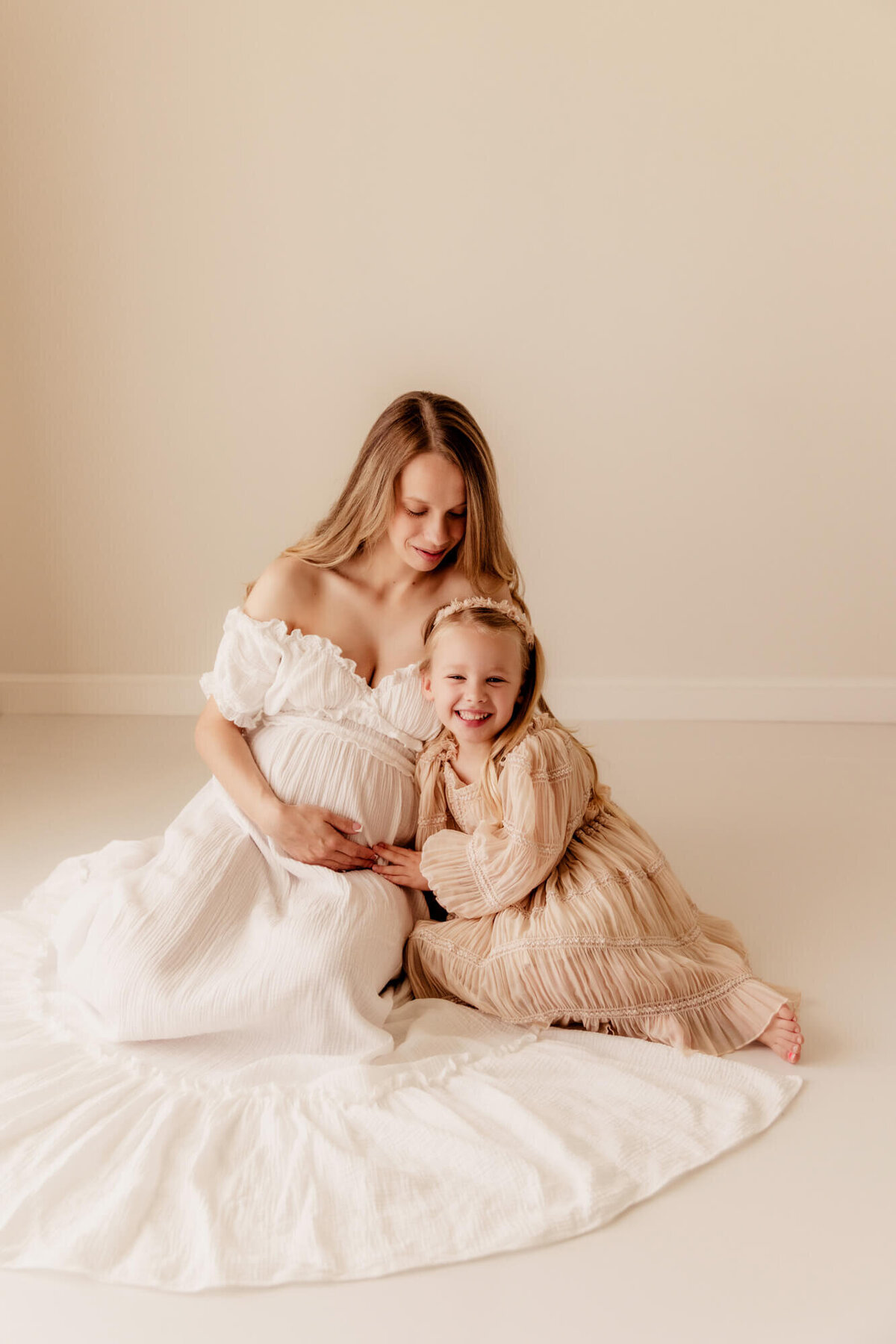 Girl laughs while sitting next to her pregnant mother who is wearing a white dress.