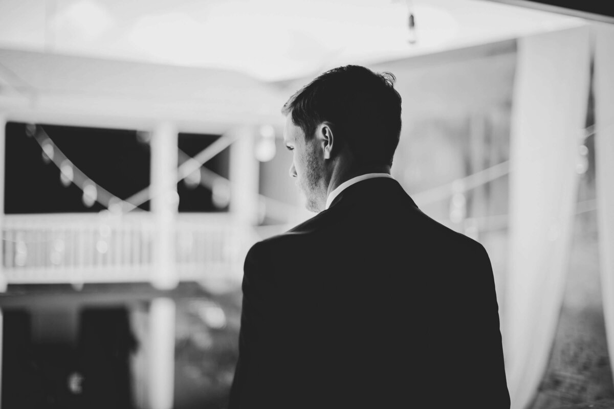 A man in a suit stands with his back to the camera, looking out over a lit balcony area at night; the image is in black and white.