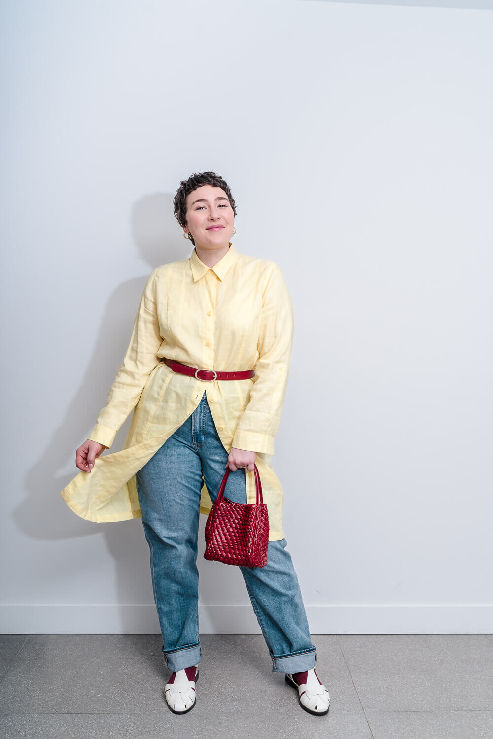 Woman in yellow shirt and jeans posing with red handbag and matching belt in front of white wall.