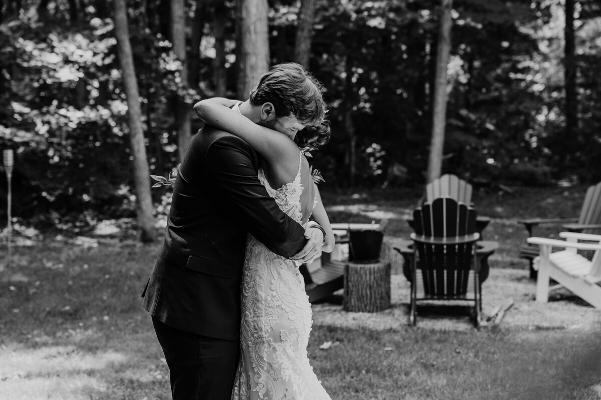 Bride and groom are hugging gently and tightly for a couples wedding day photo at Hessenland for Huron County wedding photos. Photo is captured in black and white. Behind the bride and groom is a rustic fire pit and adirondack chairs.
