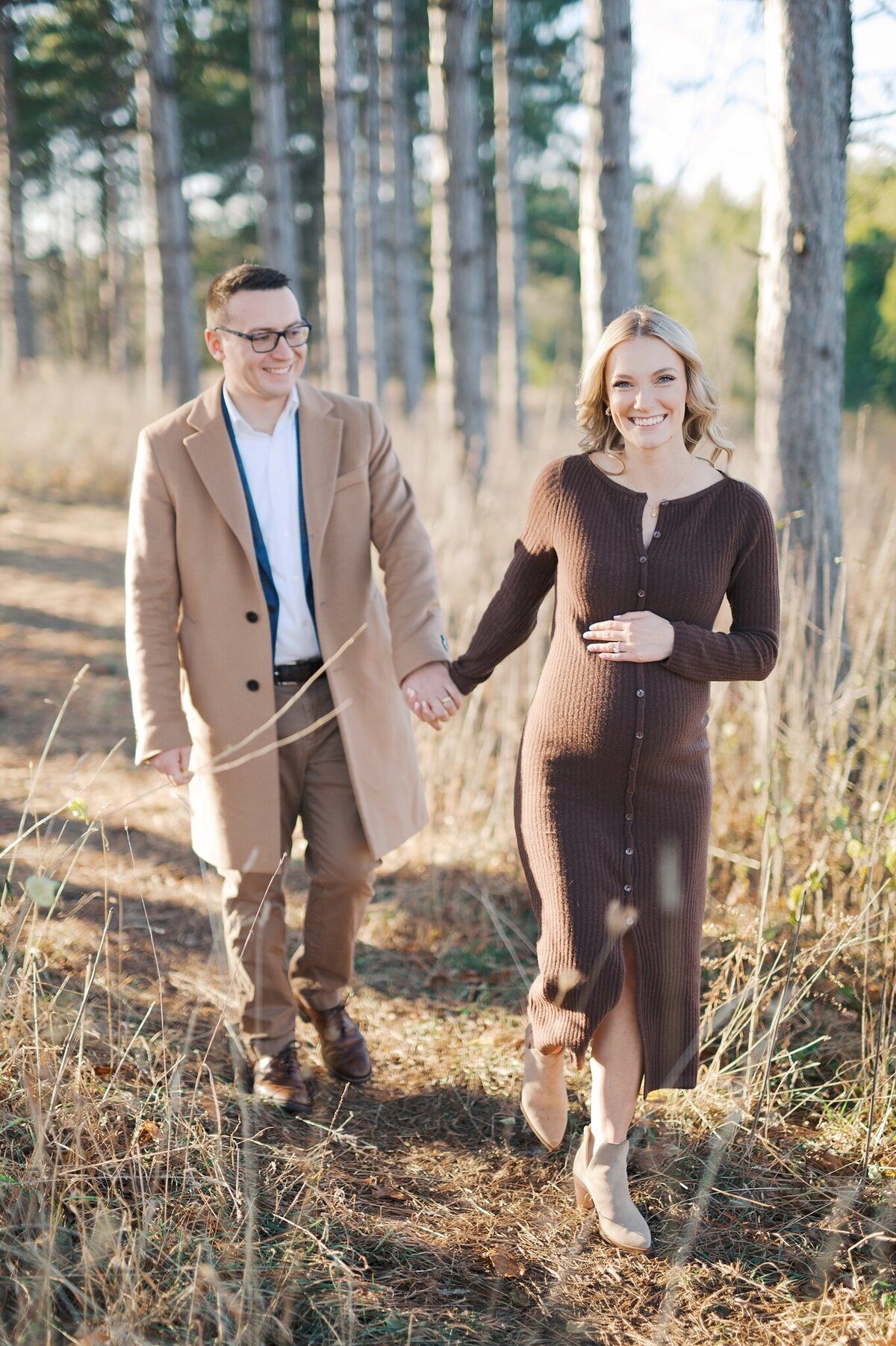 Parents-to-be holding hands and walking through pine trees in Waukesha county at Retzer Nature Center