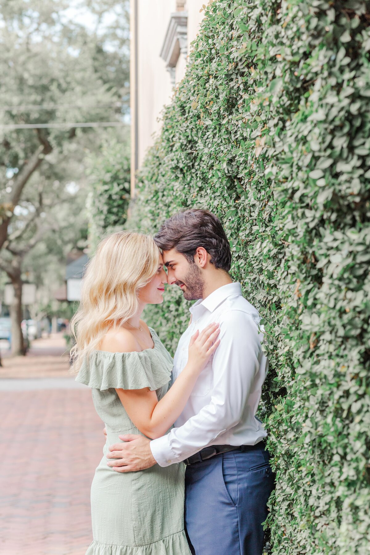 a young engaged couple embrace each other against a wall of ivy on the street.