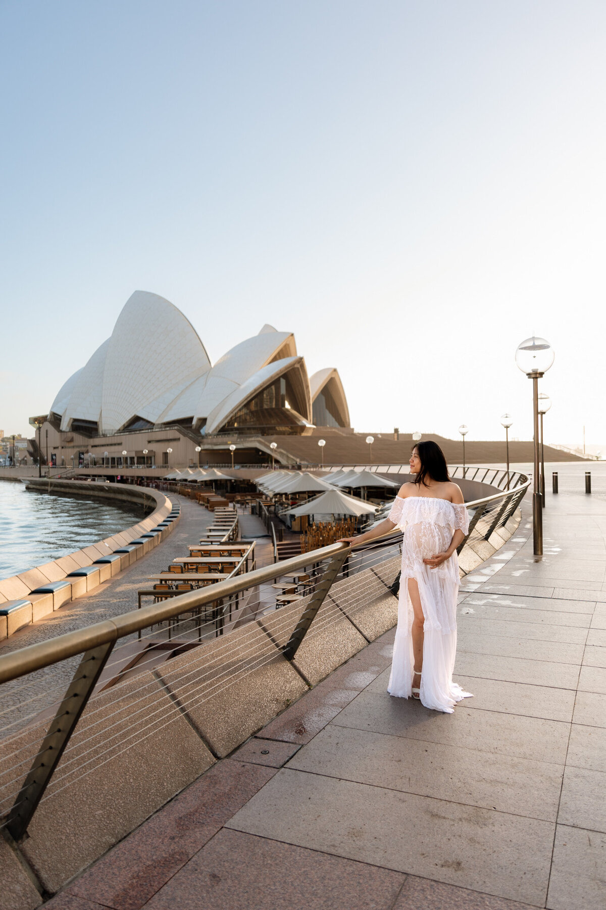 Pregnant woman in a flowing white dress standing on the Sydney Opera House promenade at sunrise, gently holding her belly and gazing toward the city skyline.
