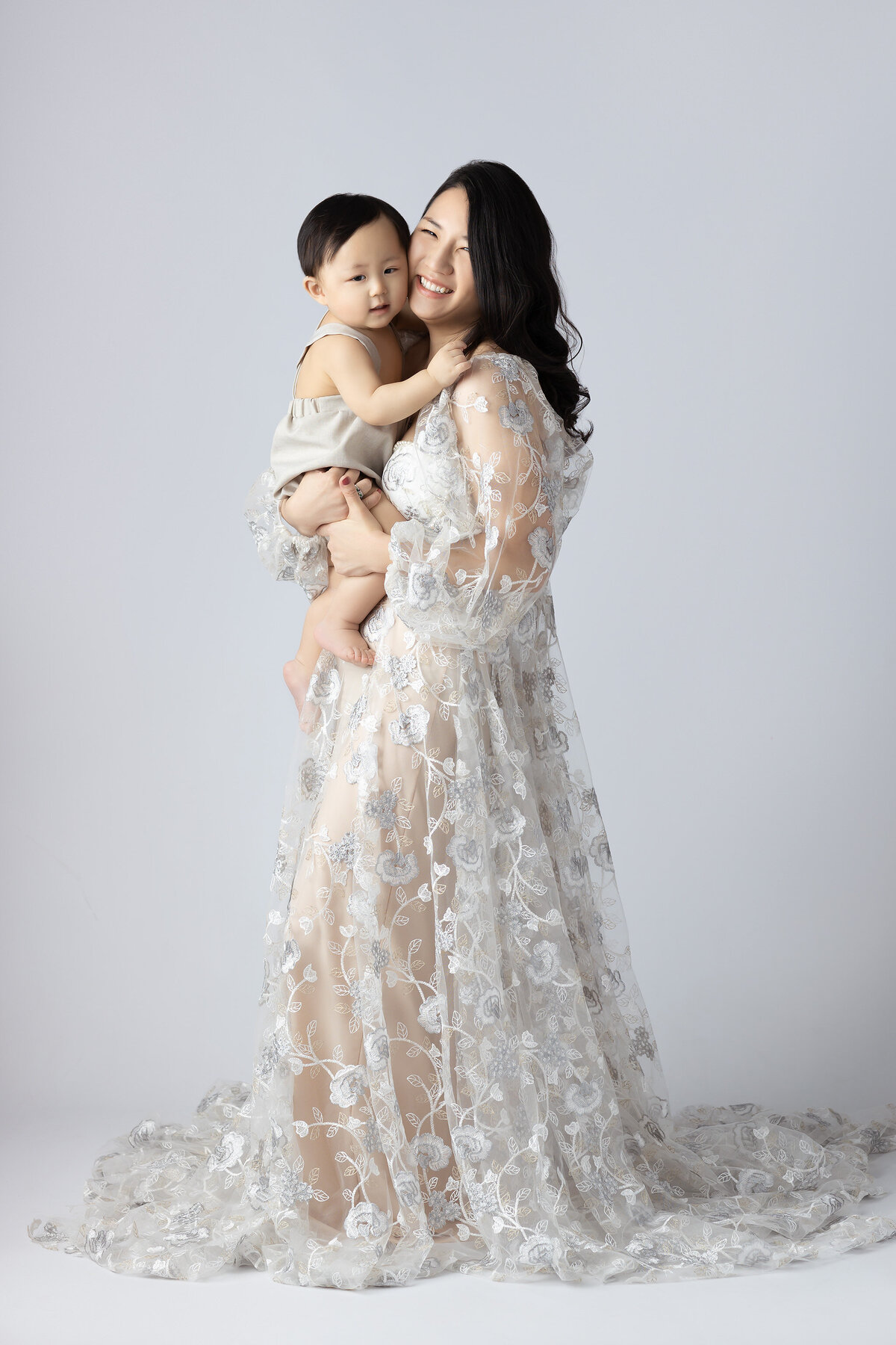 Dallas baby milestone photographer captures a joyful mother holding her son, both smiling, dressed in elegant attire, with a soft background.