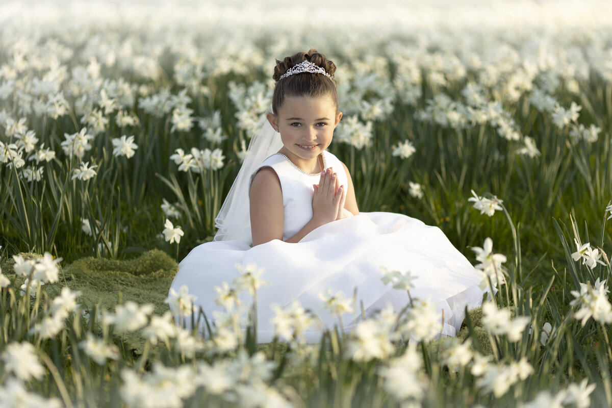 A young girl sits among white wildflowers with hands in prayer in a white dress