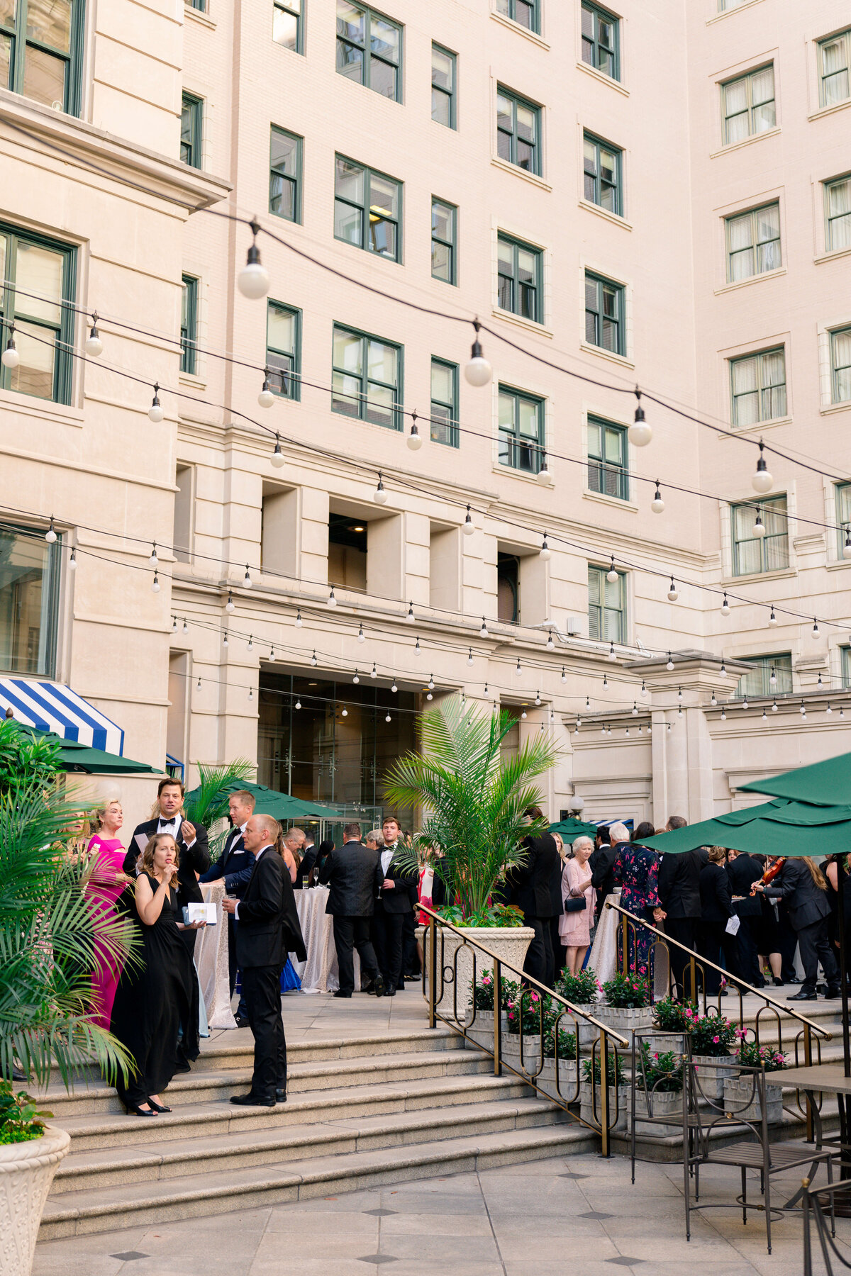 A group of people in formal attire socialize at an outdoor event. They stand on steps and a patio area adorned with string lights and potted plants. The background features a multi-story building with large windows.