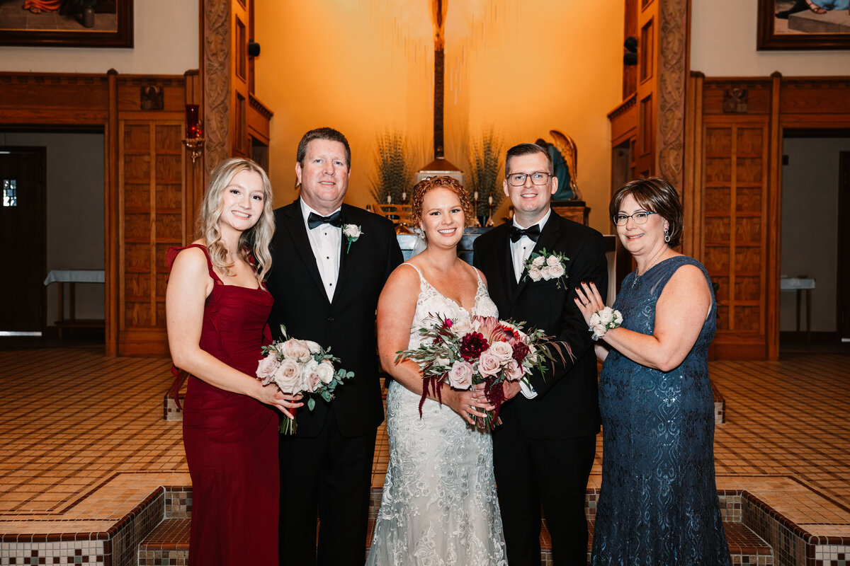 A bride and groom stand at the church altar with their family smiling