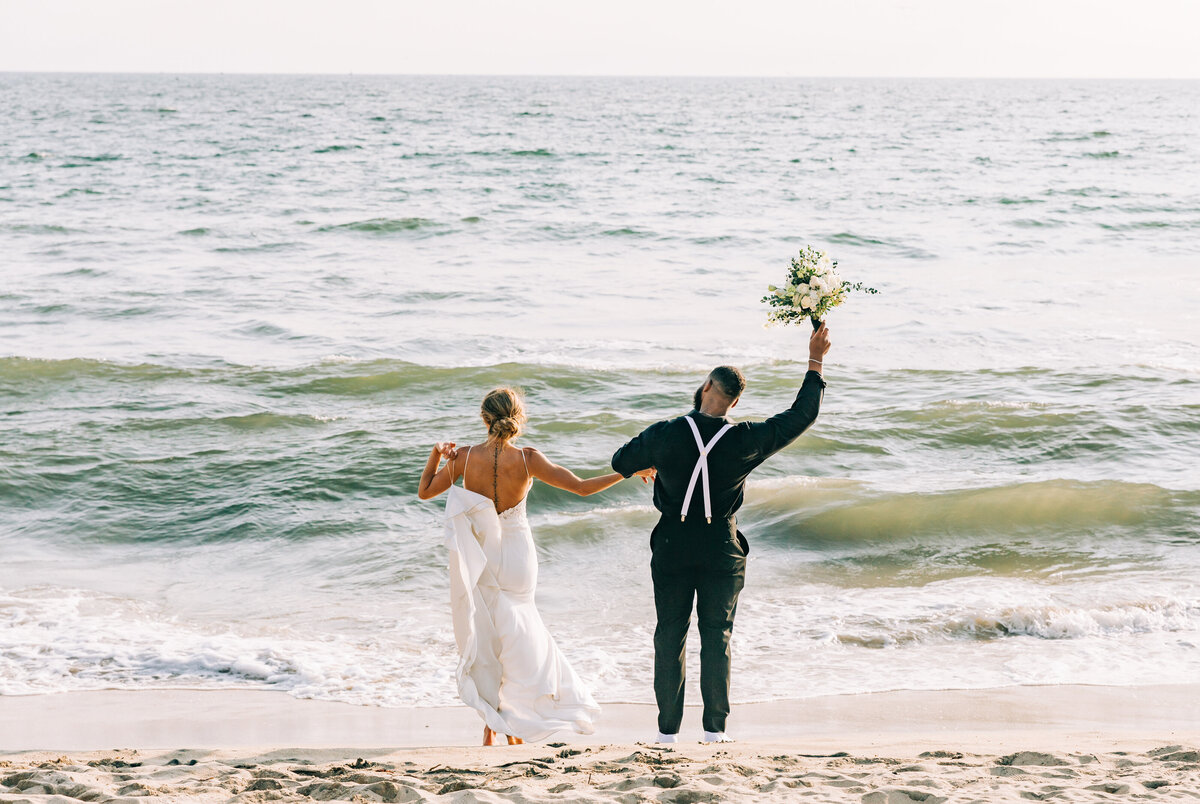 A bride and groom hold hands as they run toward the ocean on a sandy beach. The bride wears a white gown, and the groom, dressed in black, raises a bouquet into the air in celebration.