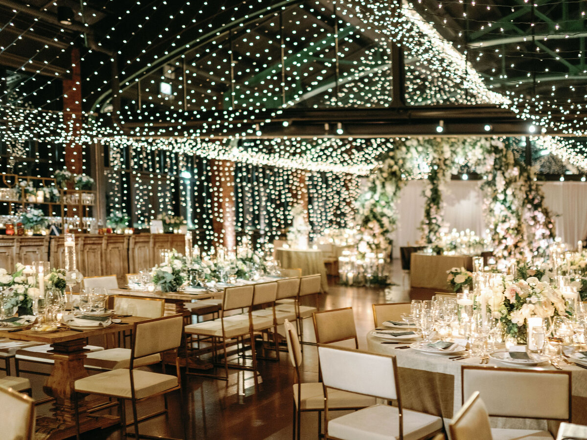 Elegant wedding reception space under a canopy of sparkling lights and greenery, with tables set in gold and white tones, ready for guests.