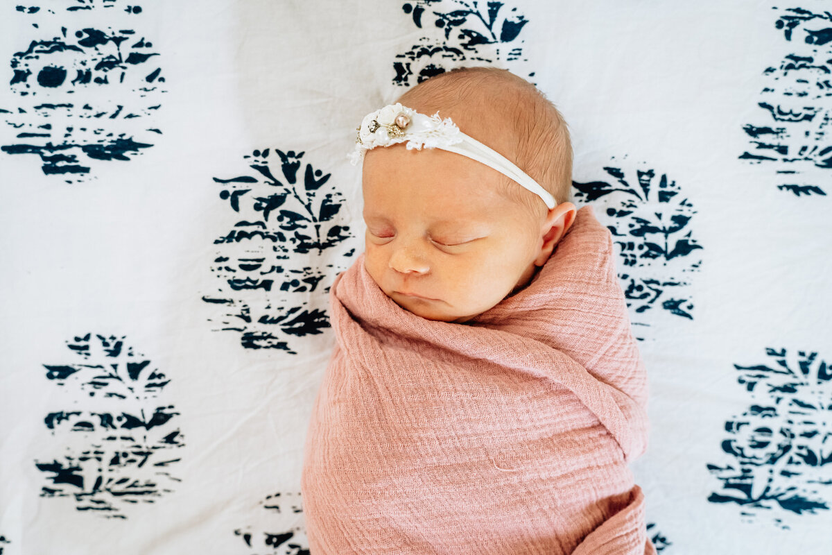A newborn baby girl wrapped in a pink swaddle, sleeping peacefully on a bed with a black and white patterned bedspread.