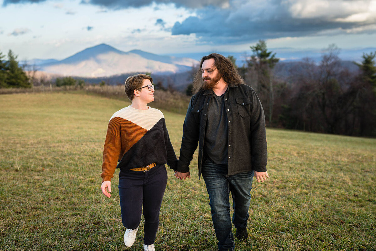 A Virginia wedding photographer holds hands and smiles  as she looks at her partner as they walk in a field with  Cahas Mountain in the background near Floyd, Virginia.