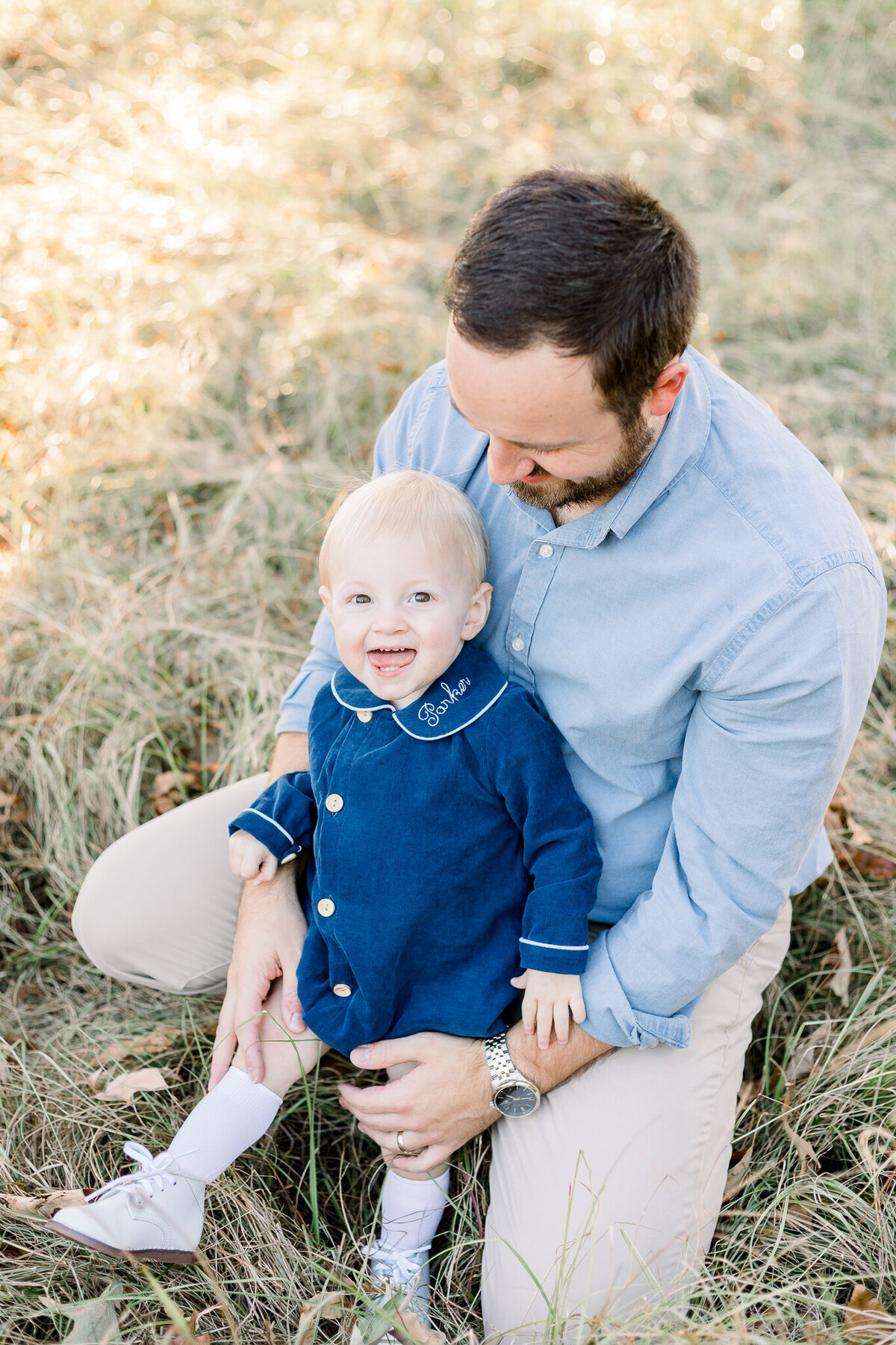 Little boy in blue smiling at camera in dad's arms in an outdoor field session by Brandon, MS Photographer.