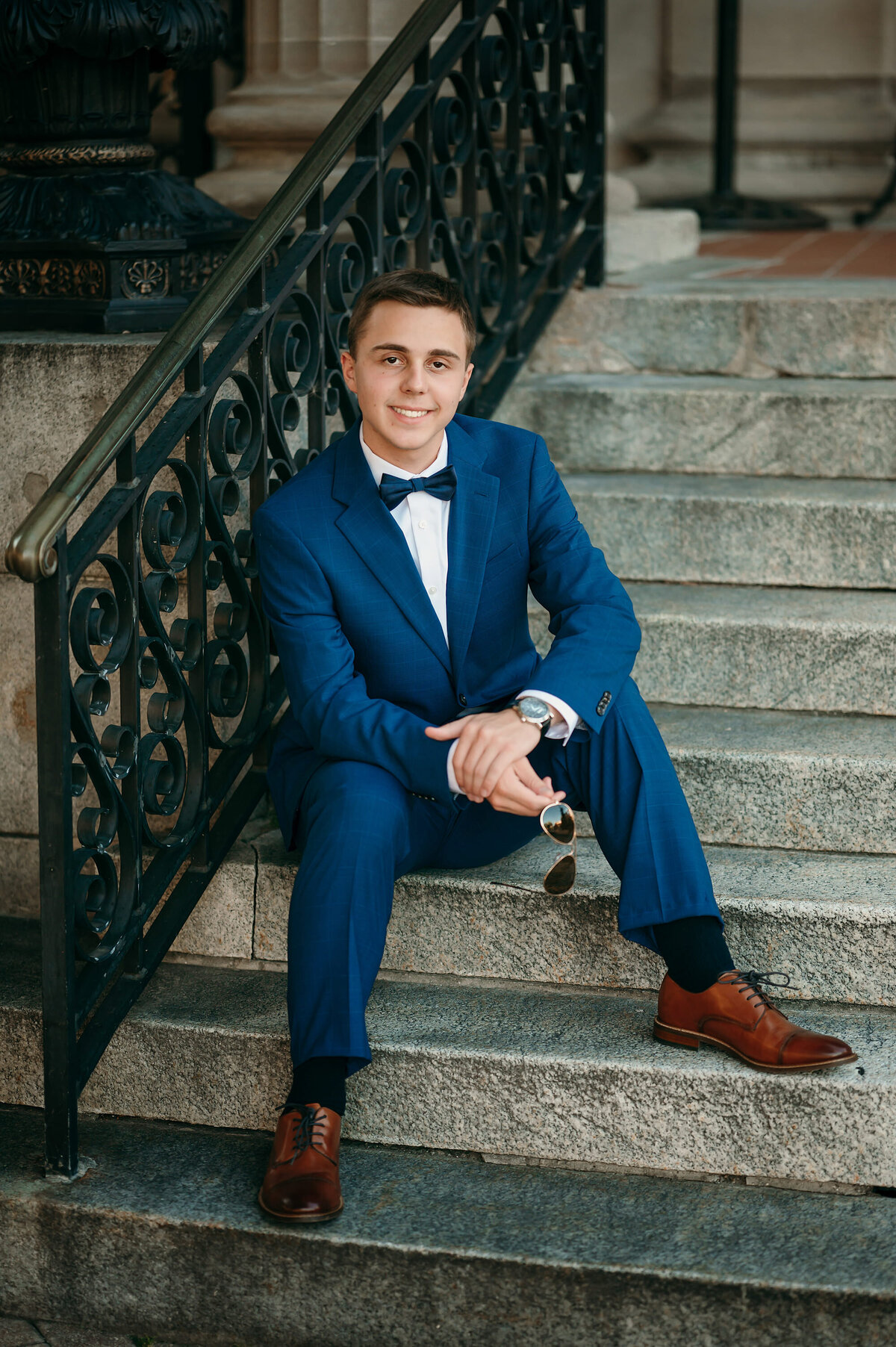 A senior student from Oconomowoc High School sits on a set of concrete steps in Hartland, WI wearing a navy suit and black bow tie.