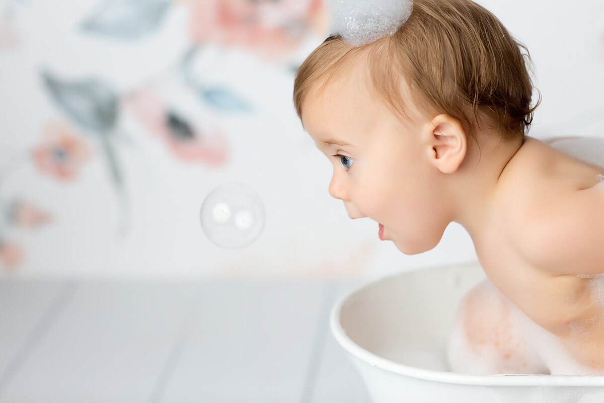 A wide-eyed baby in a white tub filled with bubbles leans forward in excitement, mesmerized by a floating bubble during a playful milestone session by a DFW milestone photographer.