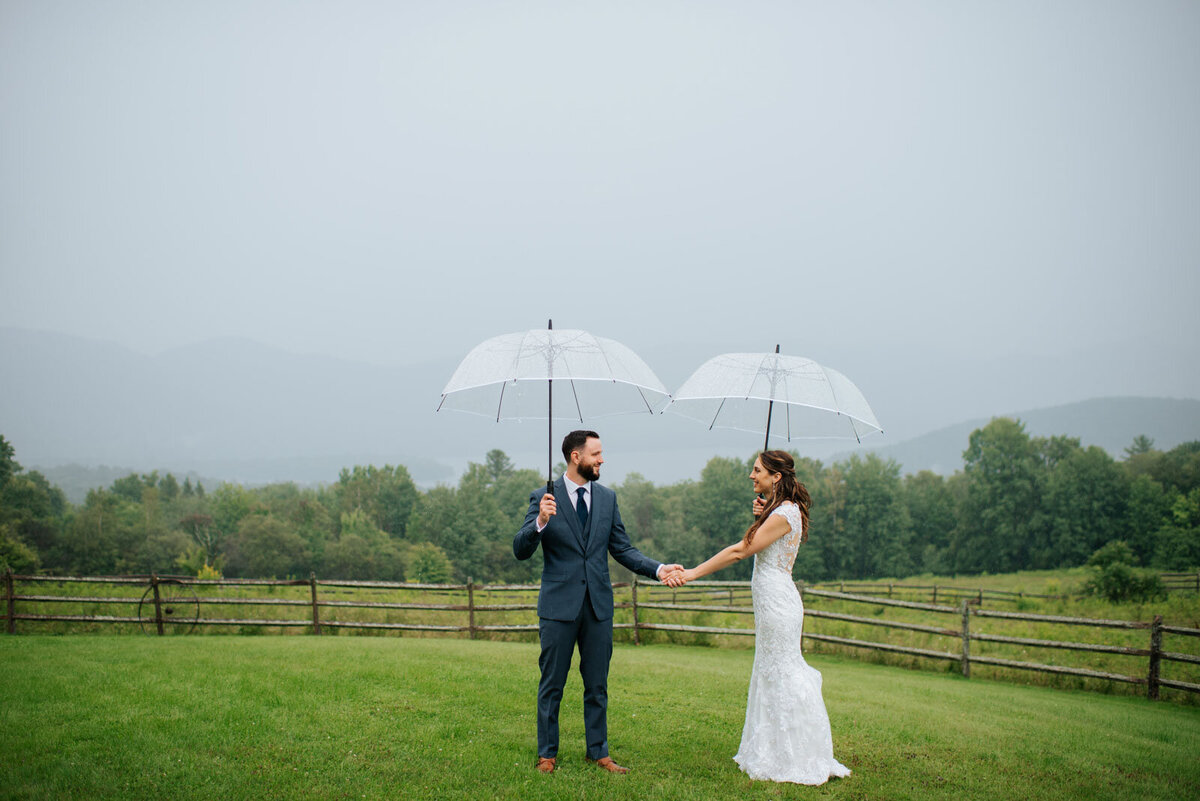 bride and groom standing in field holding umbrellas in the rain at vermont mountain top inn and resort wedding