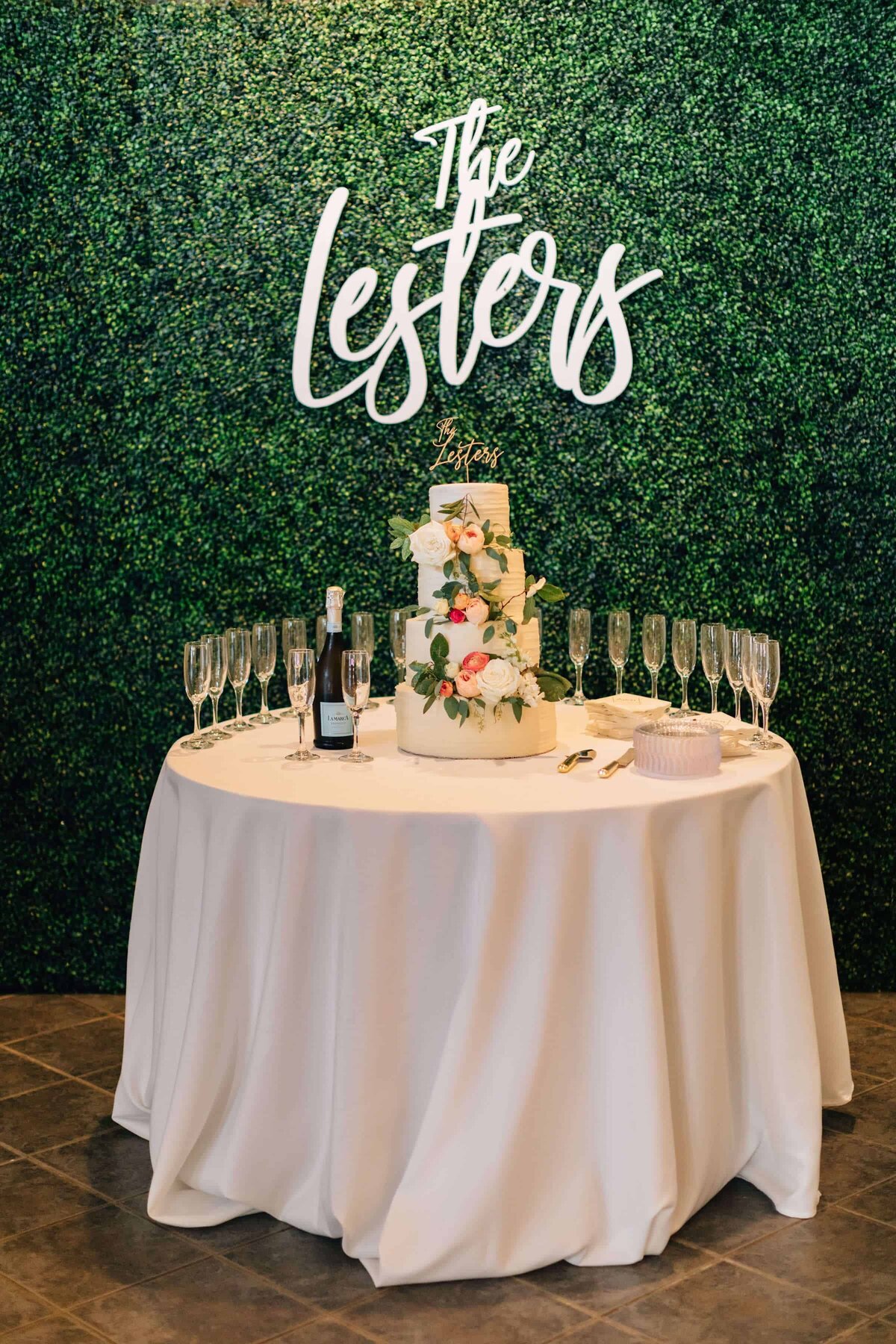 A round table with a white cloth holds a two-tiered floral cake and glasses. Behind it is a green leafy wall with The Lesters sign in white script at the top.