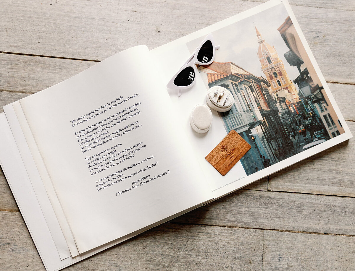 Open book with a poetic Spanish excerpt and a scenic photo of Cartagena Colombia, styled with white sunglasses, a wooden card, and wedding rings. Captured by Claudia Amalia Wedding Photography for Victoria and Daniel’s Cartagena Colombia wedding.