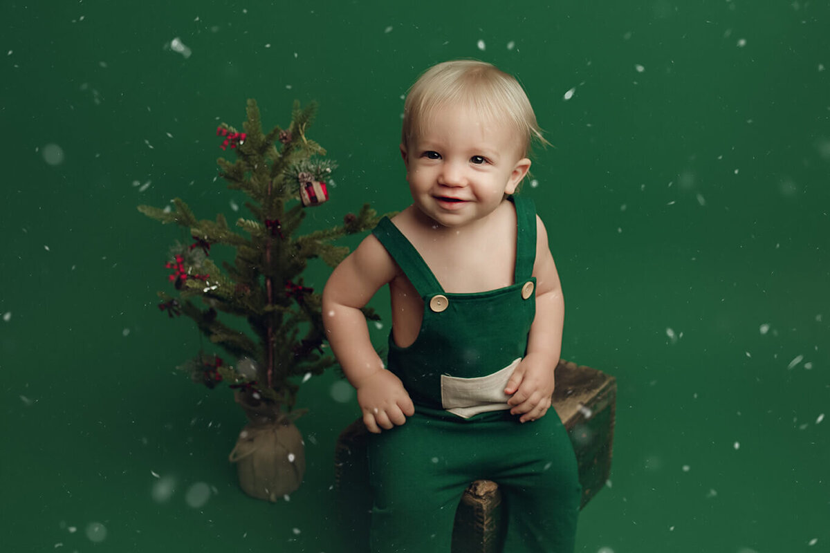 a one year old boy sitting on a green drop with a christmas tree and snow falling