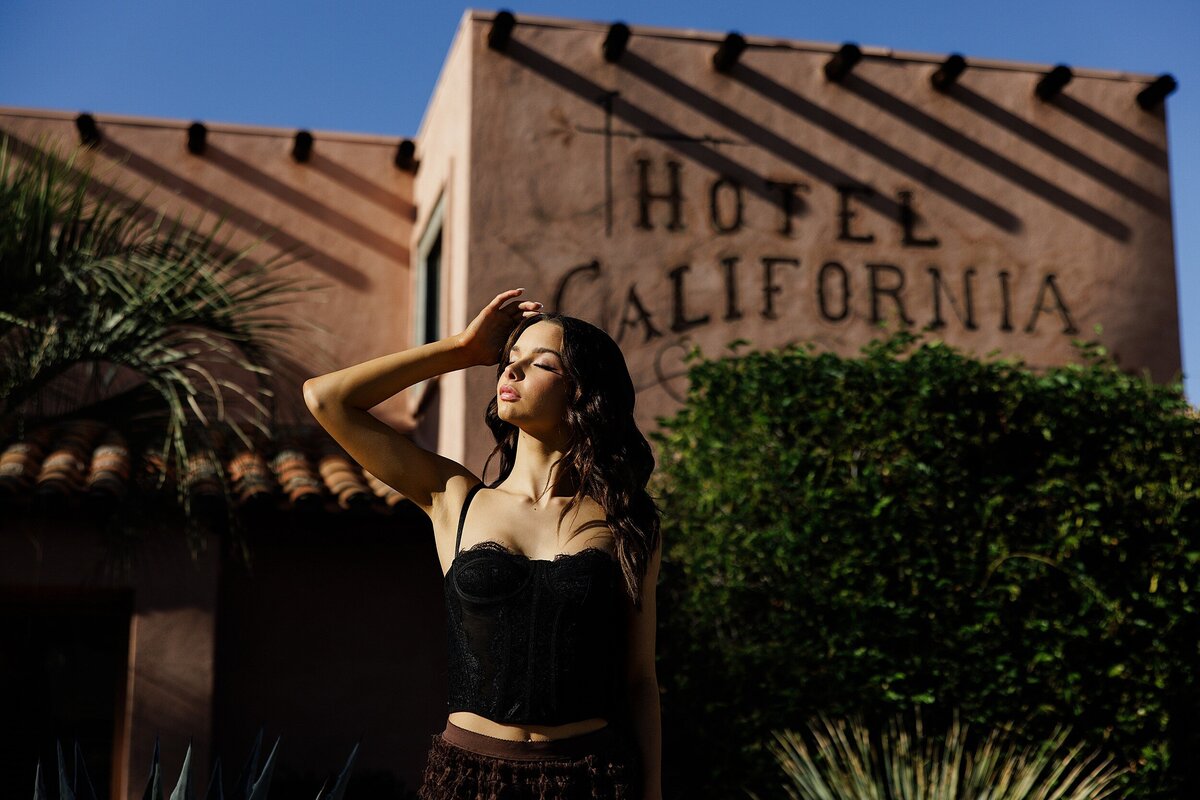Dramatic senior portrait of a girl in a black crop top shielding her eyes from the sun in front of the iconic Hotel California in Palm Springs, California, desert vibes included 