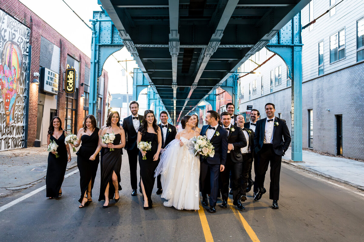 A wedding party in formal attire walks under a blue bridge structure on a city street.