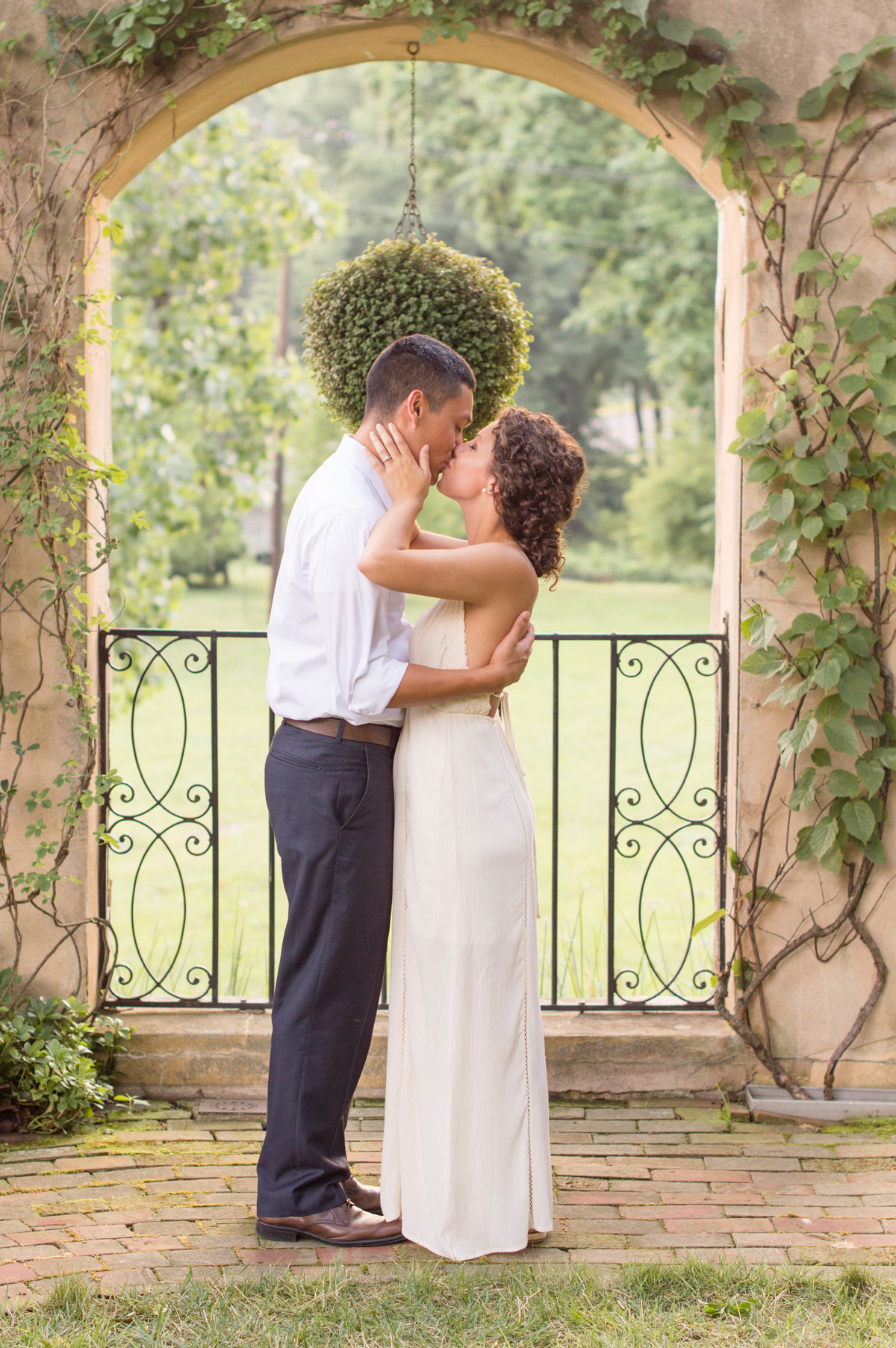 Couple hugging tightly and kissing by vine-covered archway at the Conestoga House & Gardens.