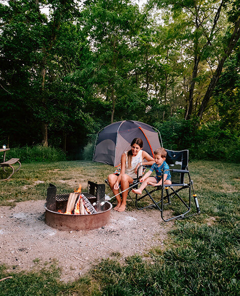 A woman and child relax in chairs by a campfire at Five Rivers Metroparks, enjoying the warmth and ambiance.