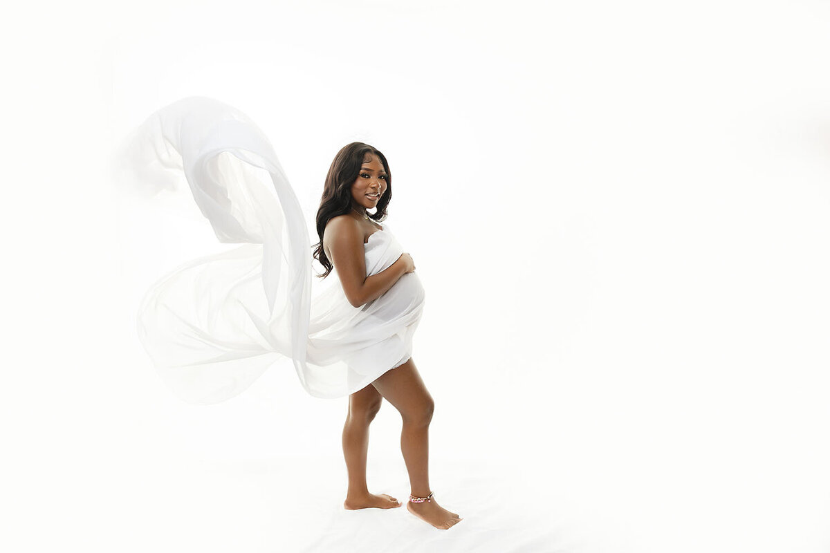 A pregnant woman stands in a white studio covering with a white sheet that flows int he wind behind her