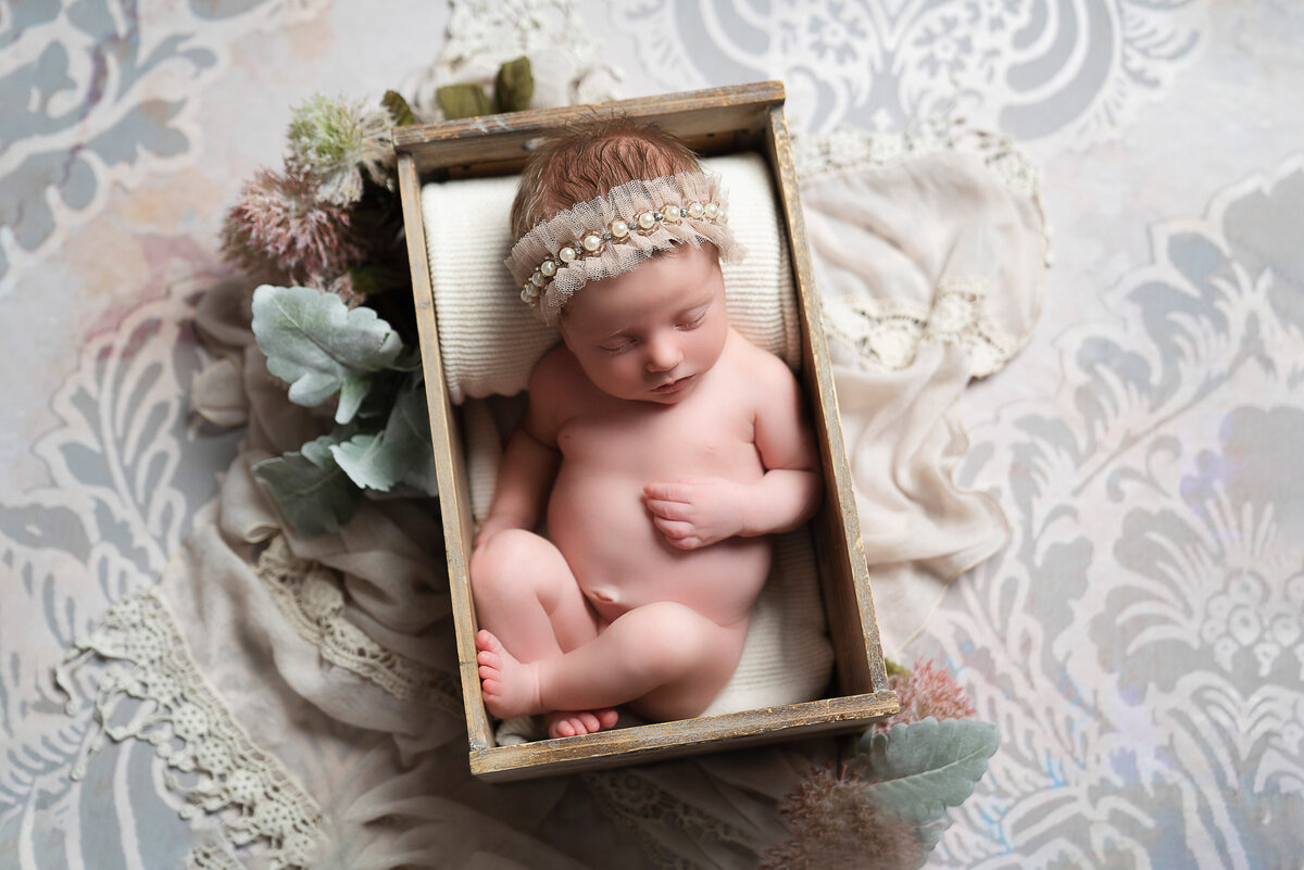 Baby girl sleeping bare for a newborn photoshoot. She is sleeping in a wooden crate atop of a damask floor drop and is wearing a beaded headband. Captured by Main Line photographer Liza Nicole.