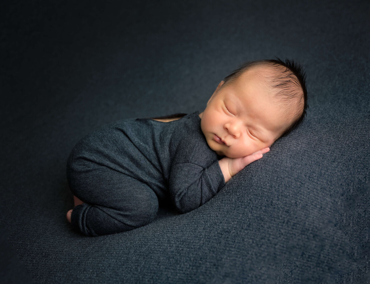 Newborn sleeping on his hands on a beanbag with dark gray cover. Image by Beth Benecki