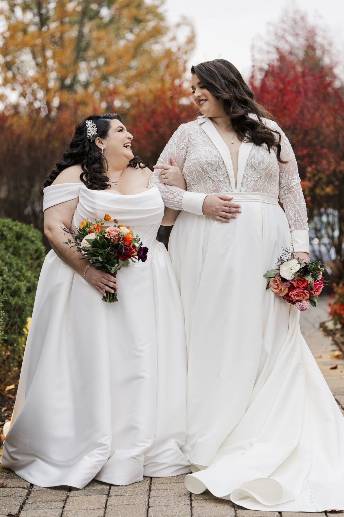 Marilena and Kristine smiling together in their wedding dresses under fall foliage