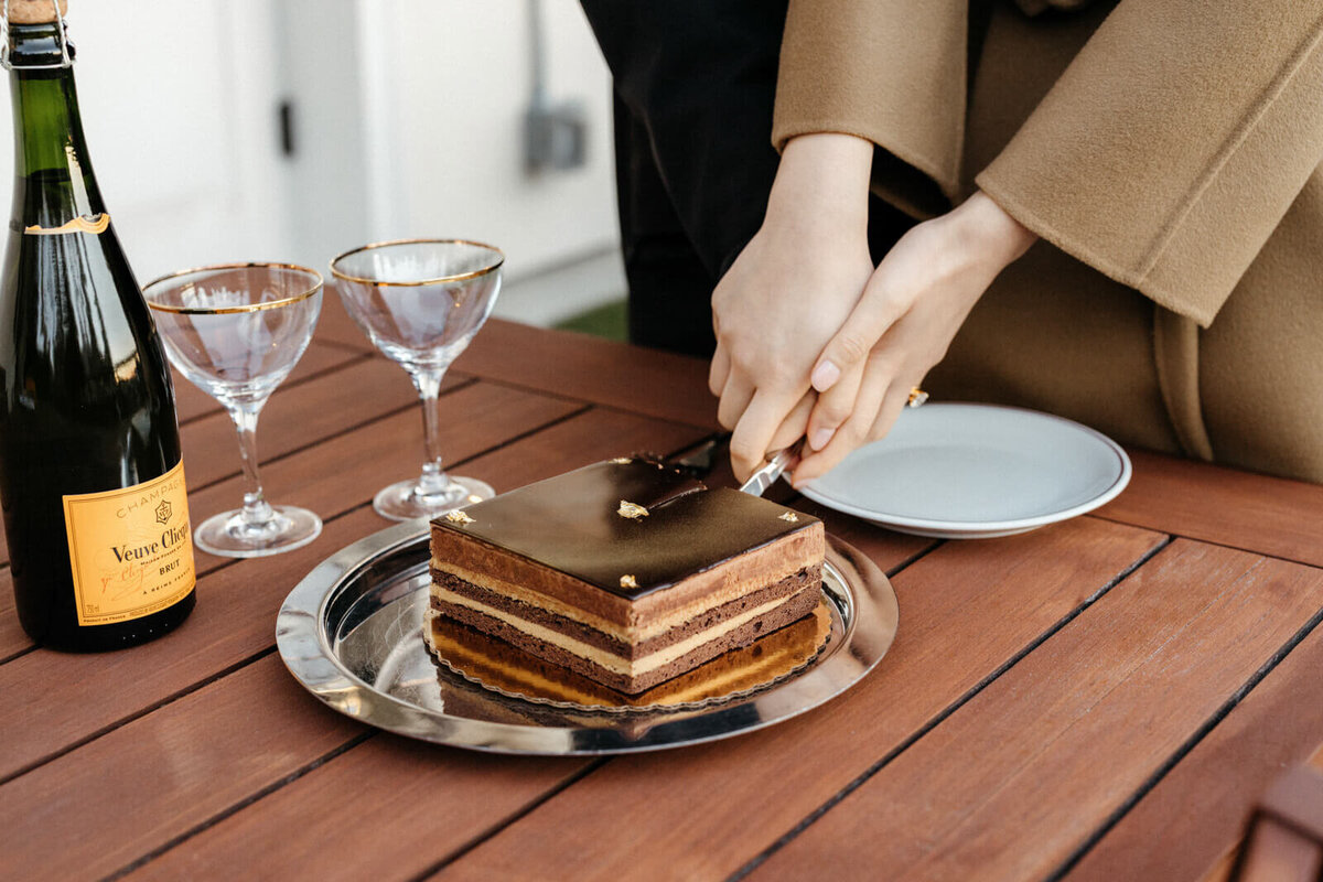 Two hands are cutting a chocolate cake on a table, with a bottle of champagne and two wine glasses on the left side.