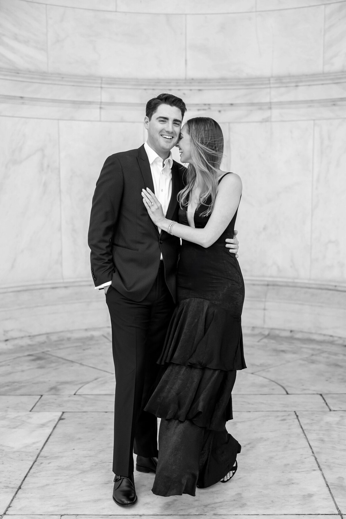 A couple stands together in an elegant setting. The man is wearing a suit, and the woman is in a long, dark dress. They are smiling and embracing, surrounded by marble walls.
