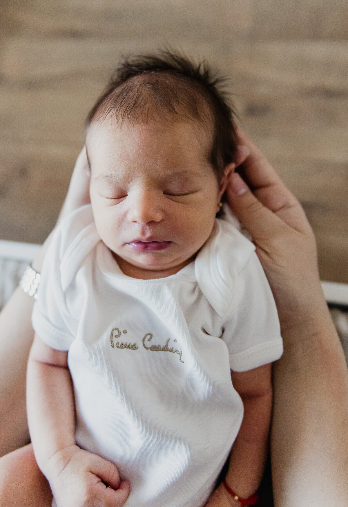 A newborn baby with dark hair sleeps peacefully while being cradled in her parents hands.