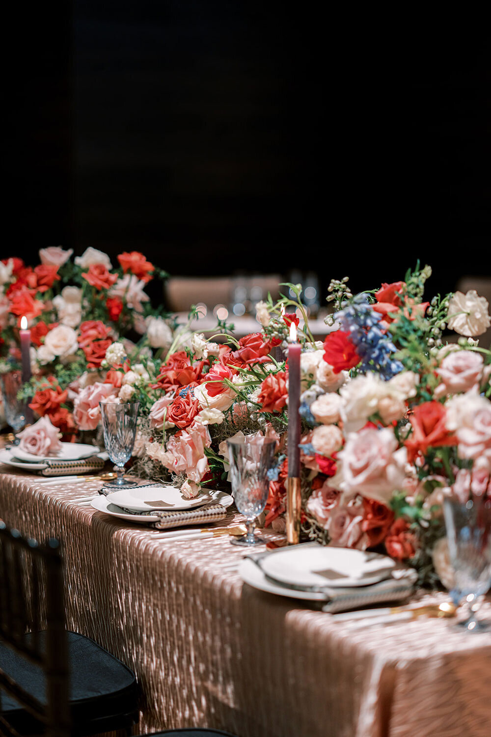 Wedding reception tablescape overflowing with red, blush, and blue florals, fine china, crystal glassware, and gold flatware atop a textured blush tablecloth.