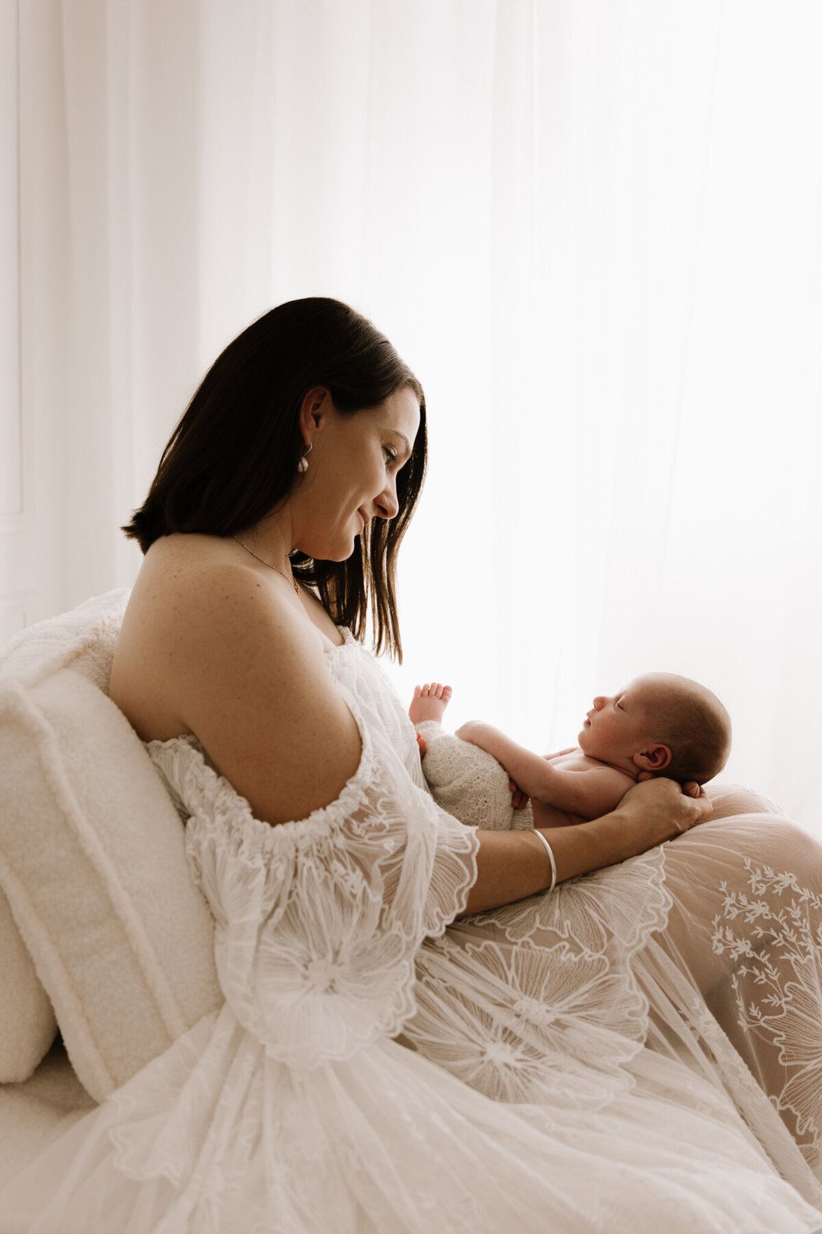 A mother in a lace gown lovingly cradling her newborn on a soft chair in a light-filled photography studio in Southwest Sydney.